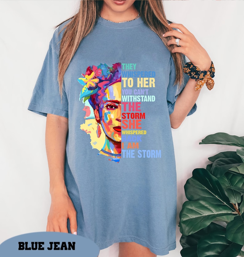 Pu&ograve; includere: Una maglietta blu con una grafica colorata del volto di una donna con accenti floreali e la scritta "THEY WHISPERED TO HER YOU CAN'T WITHSTAND THE STORM SHE WHISPERED I AM THE STORM". La maglietta ha maniche corte.