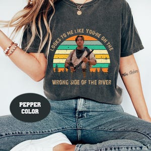 以下が含まれることがあります： 夕焼けを背景に、ライフルを持った男性のグラフィックがプリントされた、ペッパーカラーのTシャツ。「LOOKS TO ME LIKE YOU'RE ON THE WRONG SIDE OF THE RIVER」の文字が画像の上と下にプリントされています。