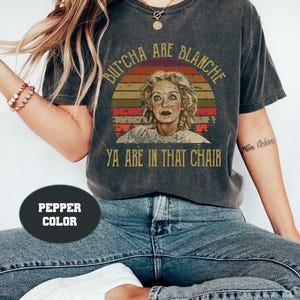 Puede incluir: Camiseta color pimienta con un gráfico del rostro de una mujer dentro de un diseño de puesta de sol. El texto de la camiseta dice "But'cha are Blanche Ya are in that chair."
