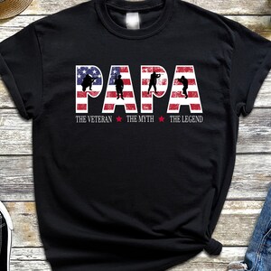 May include: Black t-shirt with a red, white, and blue American flag design. The text "PAPA" is written in white letters on the flag. Below the flag, it reads "THE VETERAN * THE MYTH * THE LEGEND".