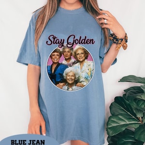 Op de afbeelding: Een lichtblauw T-shirt met een afbeelding van de Golden Girls met de tekst "Stay Golden".