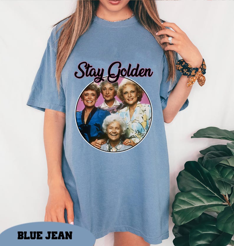 Op de afbeelding: Een lichtblauw T-shirt met een afbeelding van de Golden Girls met de tekst "Stay Golden".