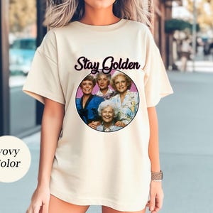 Op de afbeelding: Een wit T-shirt met een afbeelding van de Golden Girls en de tekst "Stay Golden".