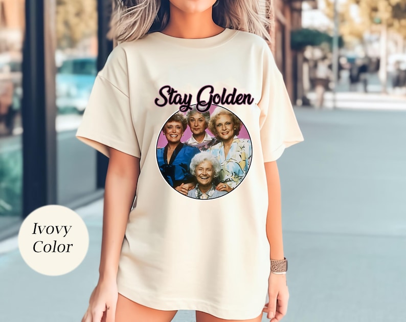 Op de afbeelding: Een wit T-shirt met een afbeelding van de Golden Girls en de tekst "Stay Golden".