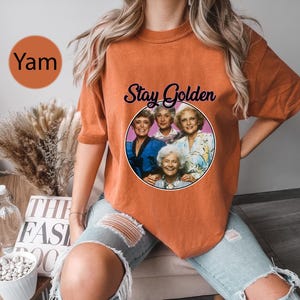 Op de afbeelding: Oranje T-shirt met een afbeelding van de Golden Girls-personages en de tekst "Stay Golden".