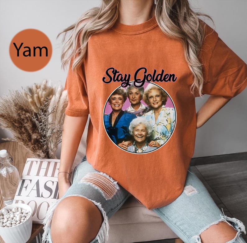 Op de afbeelding: Oranje T-shirt met een afbeelding van de Golden Girls-personages en de tekst "Stay Golden".