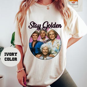 Op de afbeelding: Ivoorwit T-shirt met een afbeelding van de Golden Girls, een groep van vier vrouwen, met de tekst "Stay Golden" erboven.