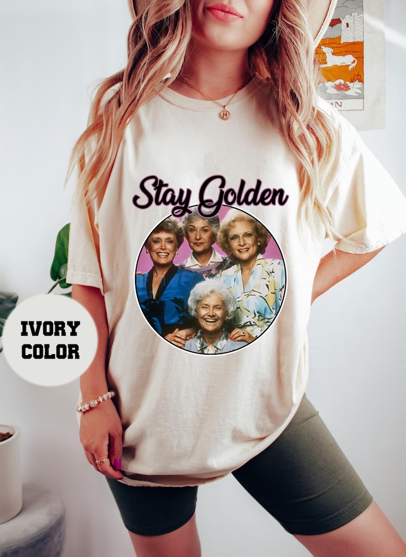 Op de afbeelding: Ivoorwit T-shirt met een afbeelding van de Golden Girls, een groep van vier vrouwen, met de tekst "Stay Golden" erboven.