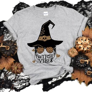 May include: A gray t-shirt with a black witch graphic wearing a leopard print hat and sunglasses. The text "Witch Vibes" is printed below the graphic.