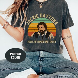 Könnte beinhalten: Ein Vintage-Style-Grafik-T-Shirt mit einem Retro-Sonnenuntergangshintergrund. Der Text "Jackie Daytona Regular Human Bartender" ist auf dem Shirt aufgedruckt.