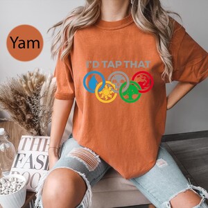 Pode incluir: Uma mulher vestindo uma camiseta laranja queimada com o texto "I'd Tap That" e cinco s&iacute;mbolos coloridos em um design circular. Os s&iacute;mbolos s&atilde;o uma gota d'&aacute;gua, uma caveira, um drag&atilde;o, um sol e uma &aacute;rvore.
