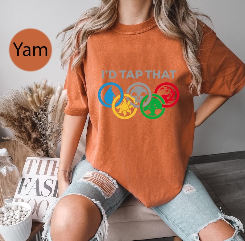 Pode incluir: Uma mulher vestindo uma camiseta laranja queimada com o texto "I'd Tap That" e cinco s&iacute;mbolos coloridos em um design circular. Os s&iacute;mbolos s&atilde;o uma gota d'&aacute;gua, uma caveira, um drag&atilde;o, um sol e uma &aacute;rvore.