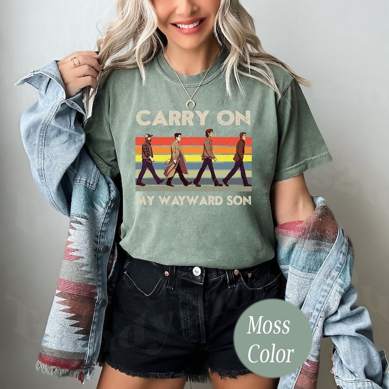 May include: A green t-shirt with a vintage-style graphic of four figures walking across a rainbow stripe. The text "Carry On My Wayward Son" is printed below the graphic. The t-shirt is a moss green color.