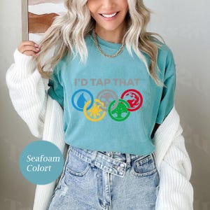 Pode incluir: Uma camiseta azul turquesa com o texto "I'd Tap That" e um gr&aacute;fico de cinco an&eacute;is ol&iacute;mpicos com s&iacute;mbolos de Magic: The Gathering em cada anel.
