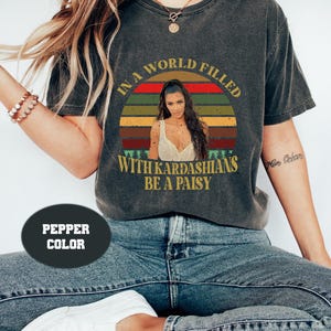 May include: Pepper-colored t-shirt with a graphic featuring a woman's image inside a sunset design. The text reads, "IN A WORLD FILLED WITH KARDASHIANS BE A PAISY."