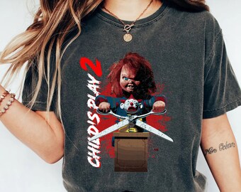 May include: A dark gray t-shirt with a graphic of Chucky from the Child's Play movie series. Chucky is holding a pair of scissors and the text "Child's Play 2" is printed on the shirt.