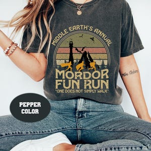 Puede incluir: Una camiseta gris oscura con un gráfico de estilo vintage de una cordillera, una torre y dragones. El texto dice "Middle Earth's Annual Mordor Fun Run" y "One Does Not Simply Walk".