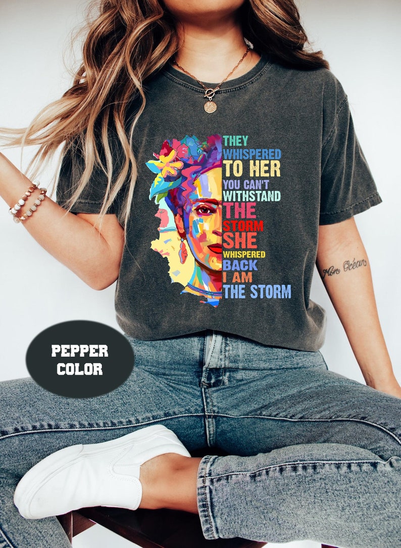 Pu&ograve; includere: T-shirt color pepe con un ritratto colorato di una donna con decorazioni floreali. La maglietta mostra il testo: "Le sussurrarono, non puoi resistere alla tempesta. Lei sussurr&ograve; di rimando, io sono la tempesta."