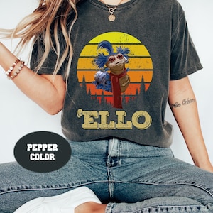 May include: A dark gray t-shirt with a vintage-style sunset graphic featuring a blue and brown creature with the text 'Ello' underneath. The shirt is labeled 'Pepper Color'.