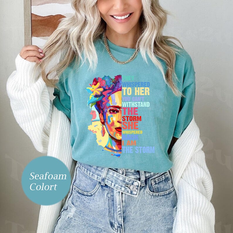 Pu&ograve; includere: T-shirt verde acqua con un ritratto colorato di una donna con fiori. La maglietta ha il testo: "Le sussurrarono, non puoi resistere alla tempesta. Lei sussurr&ograve;, io sono la tempesta."