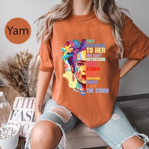 Pu&ograve; includere: T-shirt arancione a maniche corte con una grafica colorata del volto di una donna con accenti floreali. Il testo sulla maglietta recita: "THEY WHISPERED TO HER YOU CAN'T WITHSTAND THE STORM SHE WHISPERED BACK I AM THE STORM."