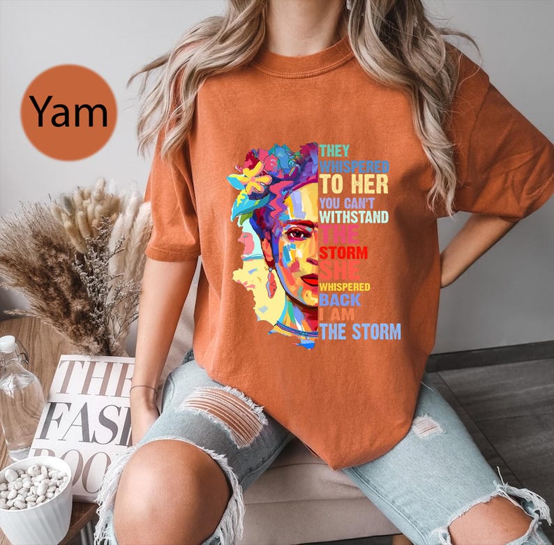 Pu&ograve; includere: T-shirt arancione a maniche corte con una grafica colorata del volto di una donna con accenti floreali. Il testo sulla maglietta recita: "THEY WHISPERED TO HER YOU CAN'T WITHSTAND THE STORM SHE WHISPERED BACK I AM THE STORM."