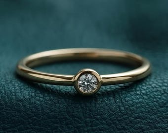 May include: A thin gold ring featuring a single, small, round, clear gemstone set in a bezel setting. The band is smooth and polished, showcasing a minimalist design with a centered stone that catches the light, creating a subtle sparkle.
