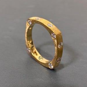 May include: A square-shaped gold ring with a textured matte finish, featuring multiple small, clear, round-cut stones embedded along the outer band. The interior of the band displays a small, circular logo with the text Rishi.
