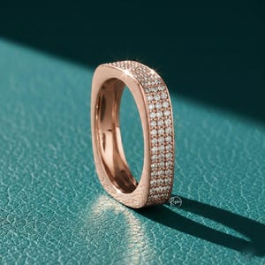 May include: Rose gold ring with a square design, encrusted with small, sparkling diamonds. The ring is set against a teal background, with a shadow cast to the right. The ring's surface reflects light, highlighting the details of the stones.