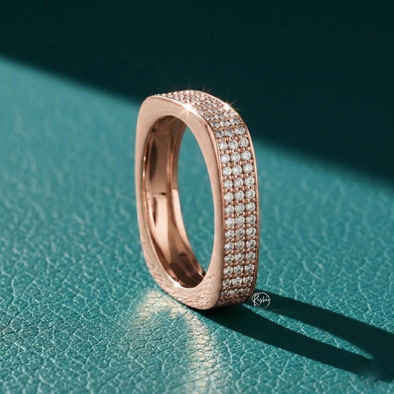 May include: Rose gold ring with a square design, encrusted with small, sparkling diamonds. The ring is set against a teal background, with a shadow cast to the right. The ring's surface reflects light, highlighting the details of the stones.