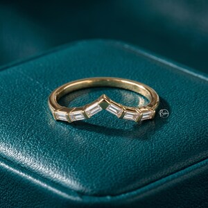 May include: A gold-colored ring with a chevron design. The ring features a series of rectangular, clear gemstones set along the top, creating a sparkling effect. The ring is displayed on a teal-colored surface.