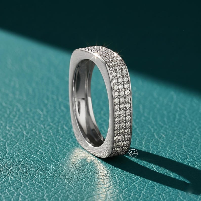 May include: A silver-colored ring with a square design, encrusted with small, sparkling stones. The ring is set against a teal-colored surface, with a shadow cast to the right. The ring's design is modern and elegant.