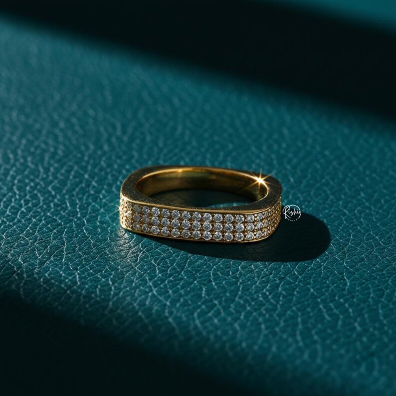 May include: A gold-toned ring with a square shape and a double row of small, clear gemstones. The ring is set against a teal-colored textured surface, with a shadow cast across the background.