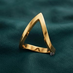 May include: A gold-toned ring featuring a distinct triangular shape with smooth, polished surfaces. The band forms a sharp point at the top, creating an open geometric silhouette. A small logo is visible near the base of the ring.