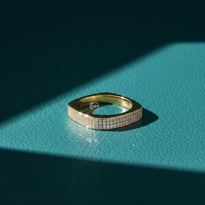 May include: A gold-colored square ring with a band of small, sparkling stones. The ring is set against a teal-colored surface, with a shadow cast across the background. The ring is a piece of jewelry.