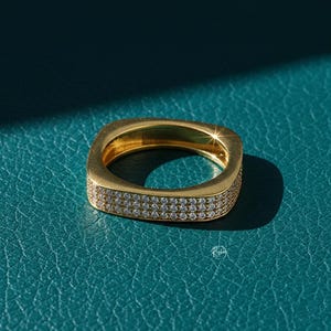 May include: A gold-colored square ring with two rows of small, clear gemstones. The ring is set against a teal-colored surface, with a shadow cast on the surface. The ring is a piece of jewelry.