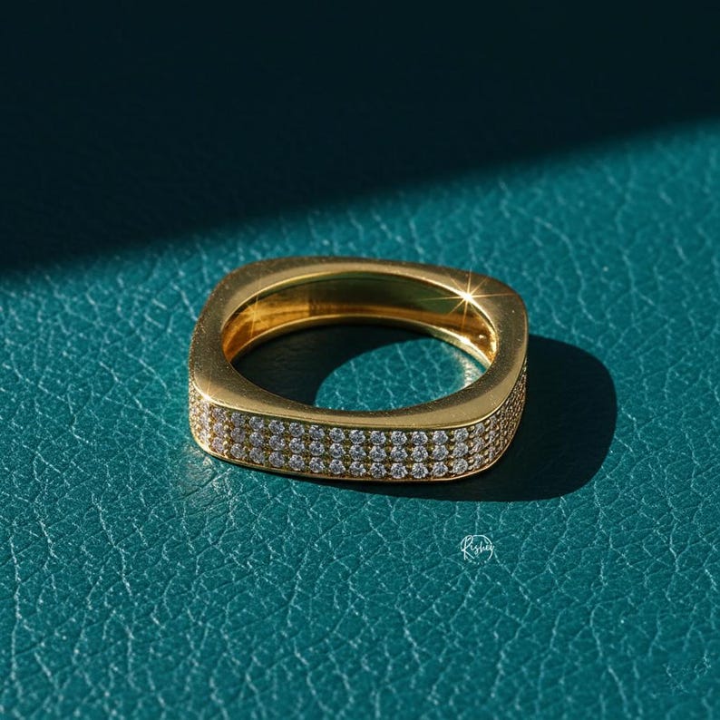 May include: A gold-colored square ring with two rows of small, clear gemstones. The ring is set against a teal-colored surface, with a shadow cast on the surface. The ring is a piece of jewelry.