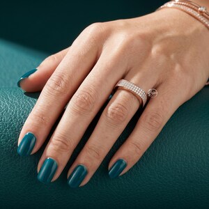 May include: Rose gold ring with two rows of small, clear stones. The ring is on a hand with teal nail polish, and a rose gold bracelet is visible in the background. The ring has a small logo on the side.
