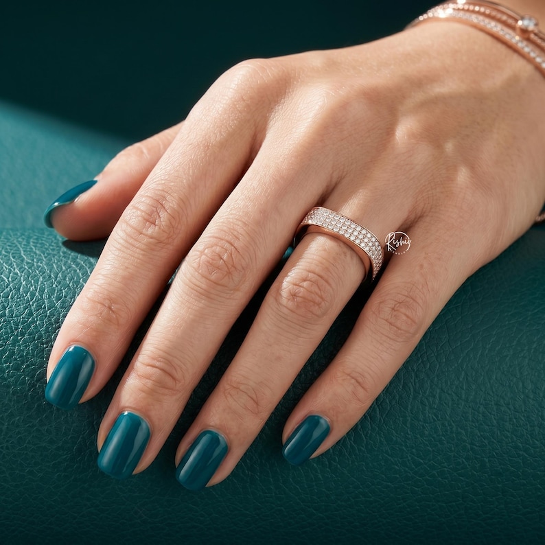 May include: Rose gold ring with two rows of small, clear stones. The ring is on a hand with teal nail polish, and a rose gold bracelet is visible in the background. The ring has a small logo on the side.