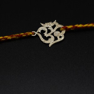 May include: A silver-colored Om symbol charm on a red and yellow braided cord. The charm is intricately designed with a detailed pattern. The cord is thick and appears to be made of woven threads, set against a black background.