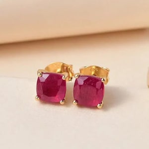 Pode incluir: Um par de brincos de ouro com pedras preciosas vermelhas escuras de corte quadrado. Os brincos são cravejados em biséis de ouro e têm pinos de ouro. As pedras preciosas têm uma aparência facetada, refletindo a luz.