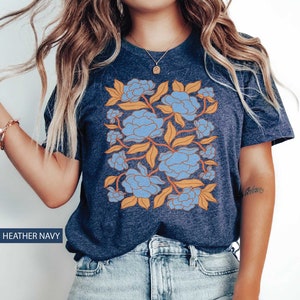Op de afbeelding: Een heideblauw t-shirt met een bloemenprint met blauwe en oranje bloemen. Het ontwerp is gecentreerd op het shirt en bedekt het grootste deel van de voorkant.