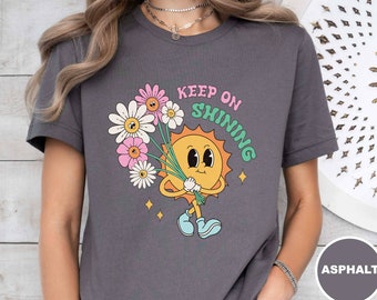 May include: A gray t-shirt with a cartoon sun holding a bouquet of flowers. The text "Keep on Shining" is printed on the shirt.