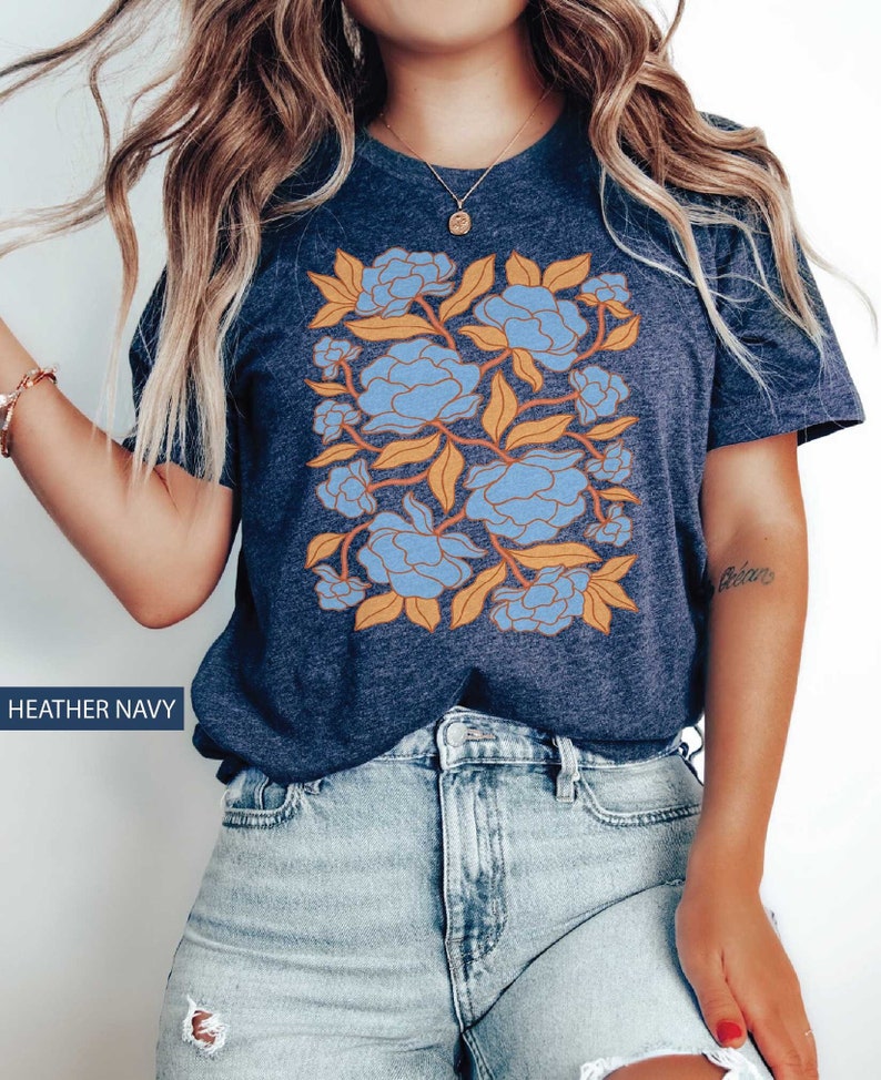 Op de afbeelding: Een heideblauw t-shirt met een bloemenprint met blauwe en oranje bloemen. Het ontwerp is gecentreerd op het shirt en bedekt het grootste deel van de voorkant.