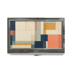 May include: A rectangular metal business card case featuring a polished silver-tone frame and a central inlay panel. The inlay displays a geometric grid pattern composed of rectangular blocks in shades of cream, navy blue, mustard yellow, and coral.