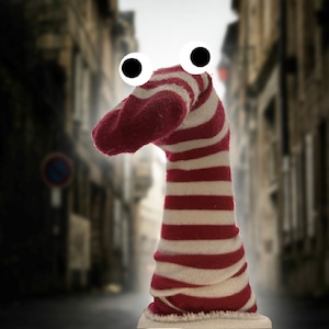Puede incluir: Un títere de calcetín a rayas rojas y blancas con grandes ojos saltones blancos. El títere está de pie frente a un fondo borroso de una calle de la ciudad.