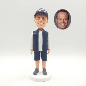 May include: A bobblehead figurine of a person wearing a navy blue vest and cap, both with "CMG" lettering. The figurine is dressed in a white shirt, navy shorts, and gray shoes. A photo of a man is in the background.