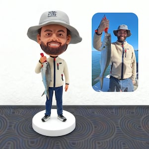 May include: A custom bobblehead figurine of a man wearing a gray hat and a cream-colored jacket, holding a fish. The figurine is on a white circular base, with a smaller image of the man holding the fish in the background.