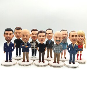 Może przedstawiać: Kolekcja spersonalizowanych figurek bobblehead, z których każda przedstawia inną osobę w różnych strojach. Figurki stoją na białych, okrągłych podstawach z imionami i tekstem. Bobbleheady mają szczegółowe rysy twarzy i są ubrane w garnitury, odzież codzienną i kostium Wonder Woman.