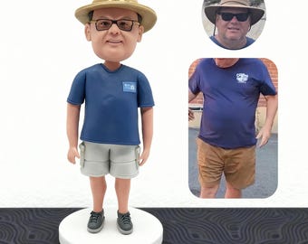 May include: A custom bobblehead figurine of a man wearing a tan hat, glasses, and a blue t-shirt with a small patch. He is also wearing gray shorts and gray sneakers. The figurine is on a white circular base.